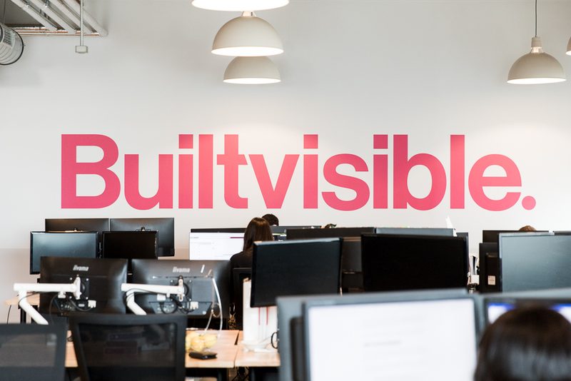 The Builtvisible office, with the pink Builtvisible wordmark on the wall and the team at their desks